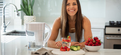 Une femme souriante dans la cuisine devant un pichet d'eau alcaline Santevia MINA blanc et des fruits frais.