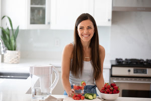 Une femme souriante dans une cuisine avec un verre d'eau minérale alcaline Santevia infusée à la fraise et au concombre provenant de son pichet MINA Santevia.