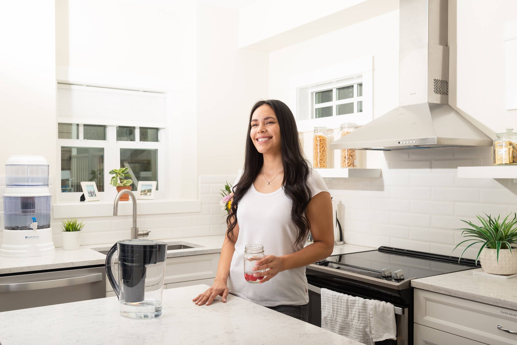 a smiling woman with brown hair in a kitchen with a Santevia Gravity Water System and a black Santevia MINA Alkaline Pitcher