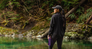 A woman staring off into the distance beside a forested lake, holding a purple Santevia Tritan Water Bottle
