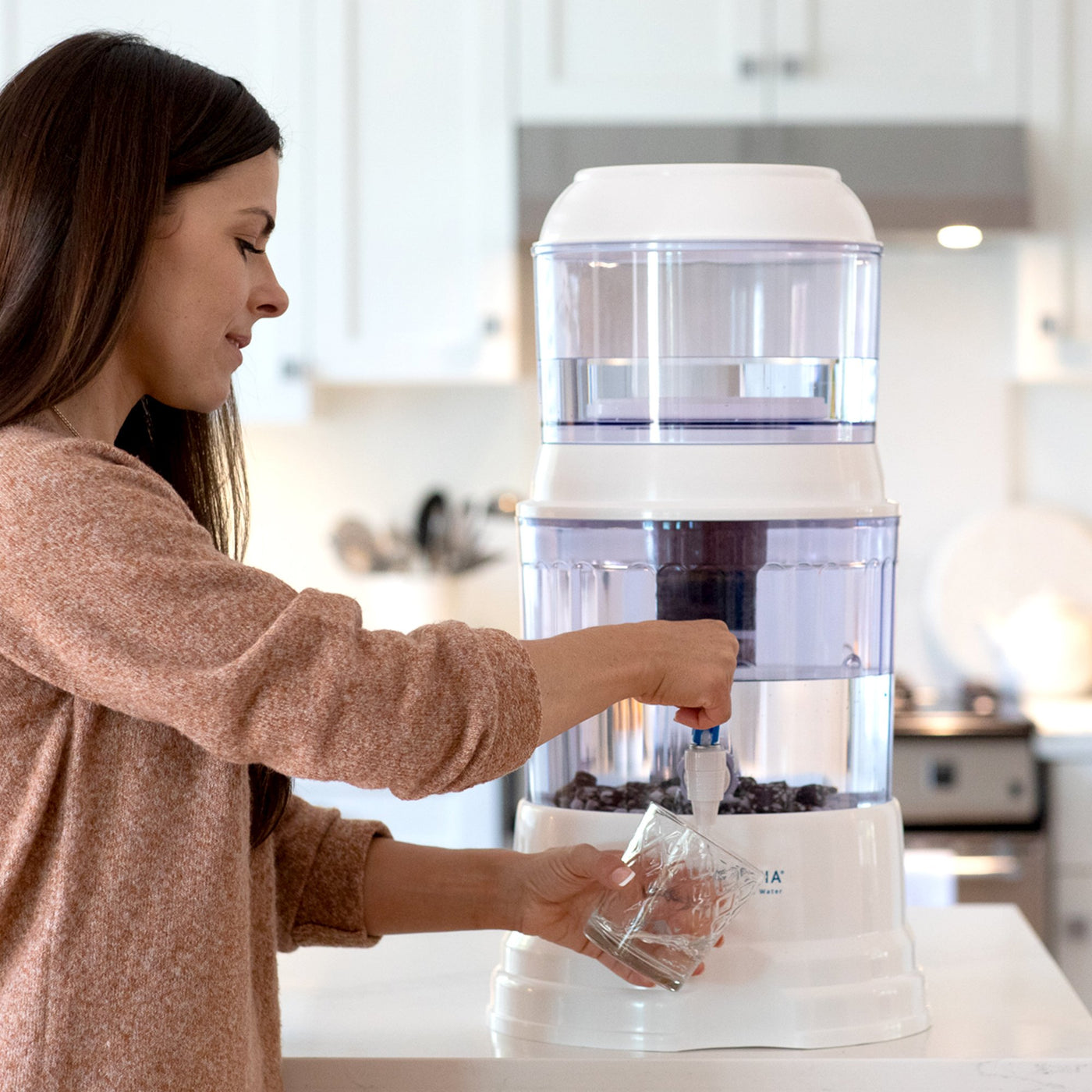  The Santevia Gravity Water System Countertop Model being used in a kitchen by a smiling woman